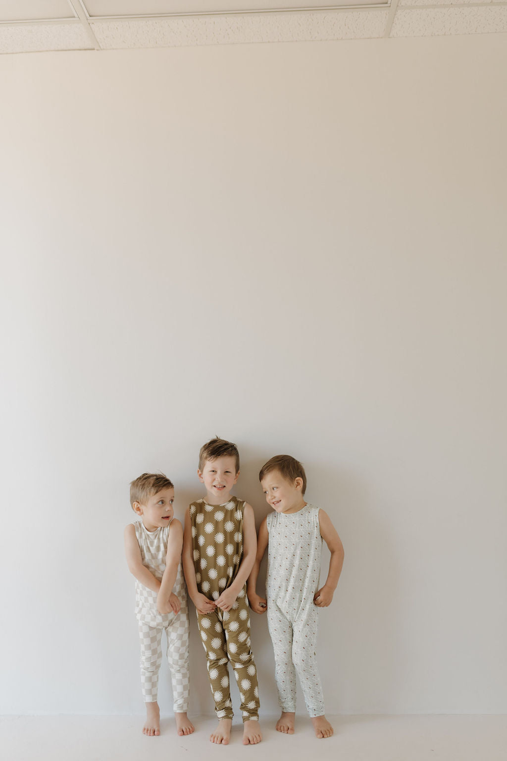 Three kids stand barefoot against a plain wall, carefree in sleeveless, neutral-toned outfits. The middle child wears a Forever French Baby Harem Bamboo Romper with brown polka dots; the others sport lighter patterns.