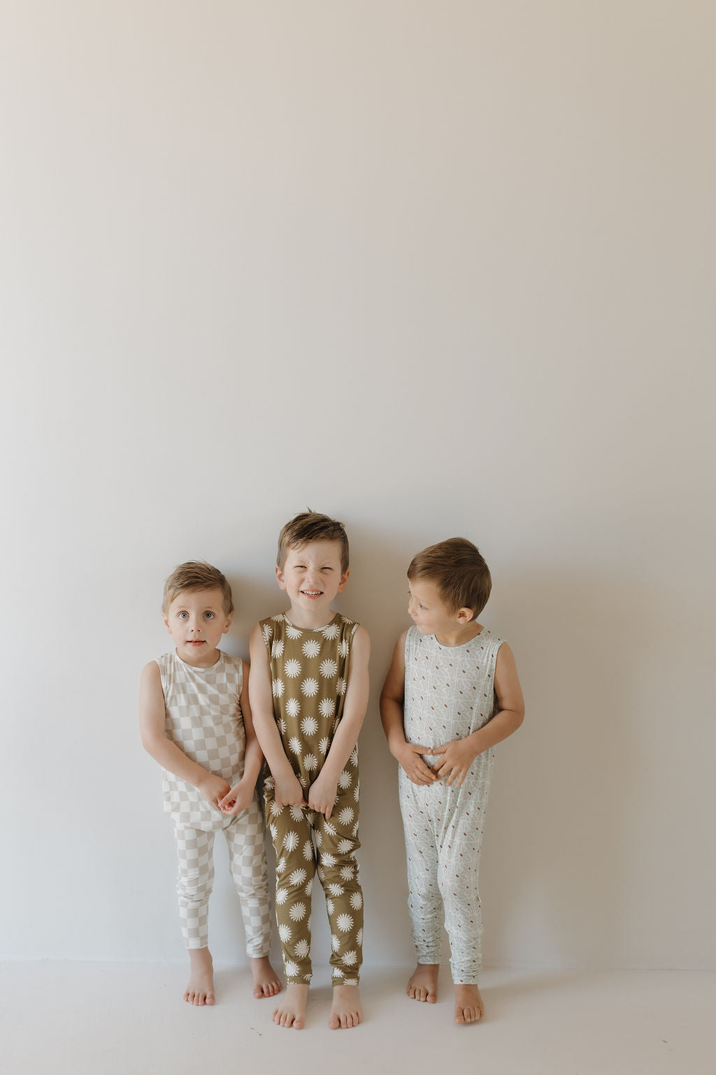 Three children stand in a row against a plain background, wearing forever french baby's coordinating sleeveless Harem Bamboo Rompers in checkered, dotted, and textured patterns. Two glance at each other while the center child smiles at the camera, showcasing their hypo-allergenic charm.
