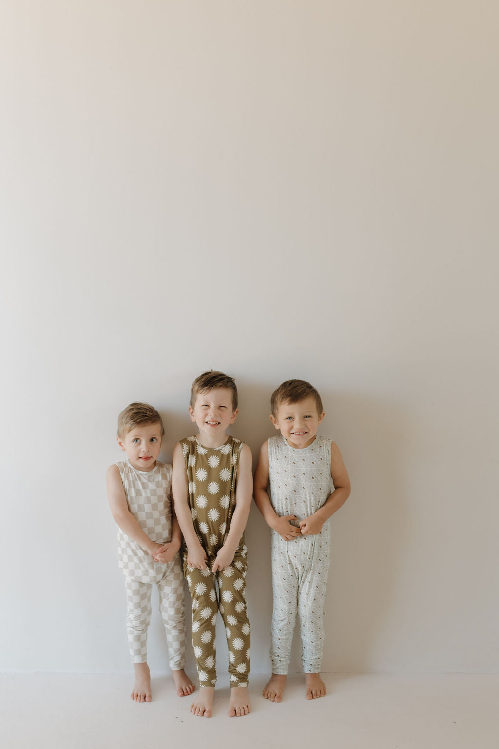 Three young children stand barefoot against a plain backdrop, smiling and wearing forever french baby's Harem Bamboo Rompers: one in white and gray checks, another in green with white polka dots, and the third in white with light prints.