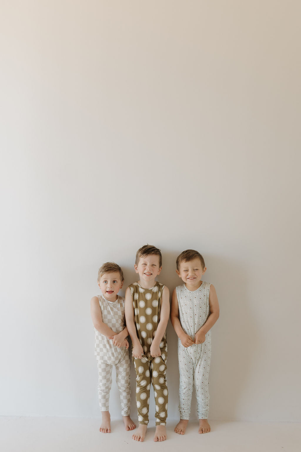 Three young children smile in sleeveless Harem Bamboo Rompers by forever french baby, standing against a white wall. Each wears a different color: beige, olive, and blue polka dots. The breathable fabric keeps them comfy as they play barefoot.