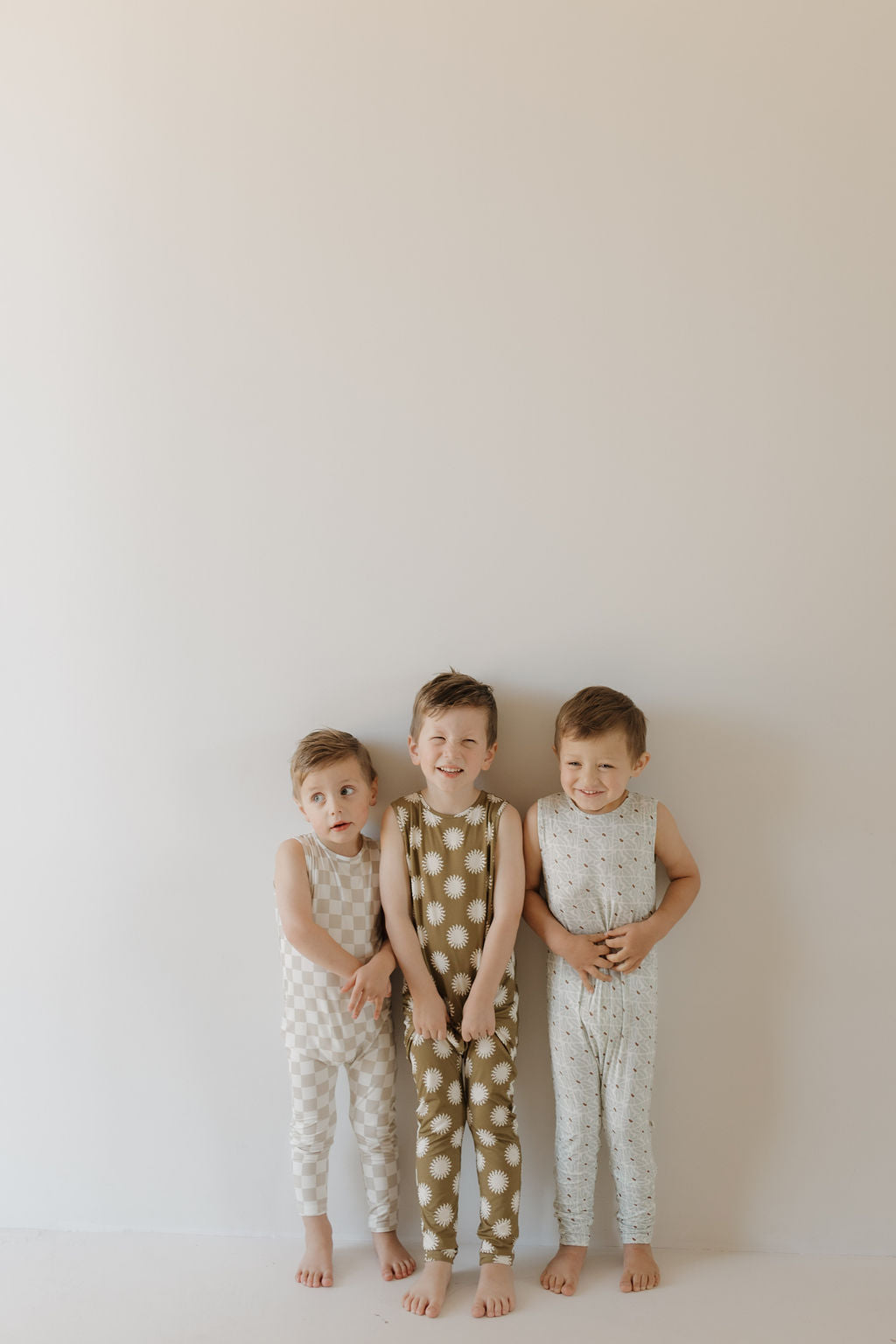 Three children smile side by side in sleeveless, hypo-allergenic Harem Bamboo Rompers. The middle child wears a floral design, while the others feature subtle patterns, all from forever french baby and made for comfort.