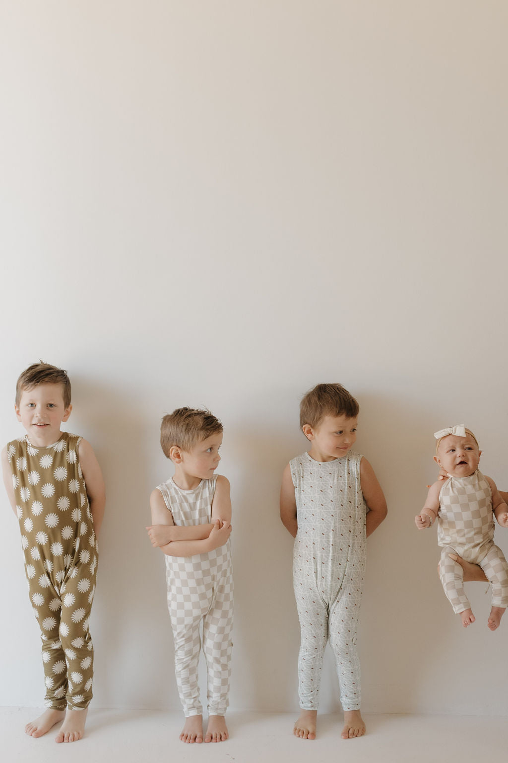 Four young children stand against a white wall wearing unique jumpsuits by forever french baby, crafted from breathable fabric: one in green with white stars, another in checkered white, one in light blue, and a baby held up wearing the beige Harem Bamboo Romper | Sol with a bow.