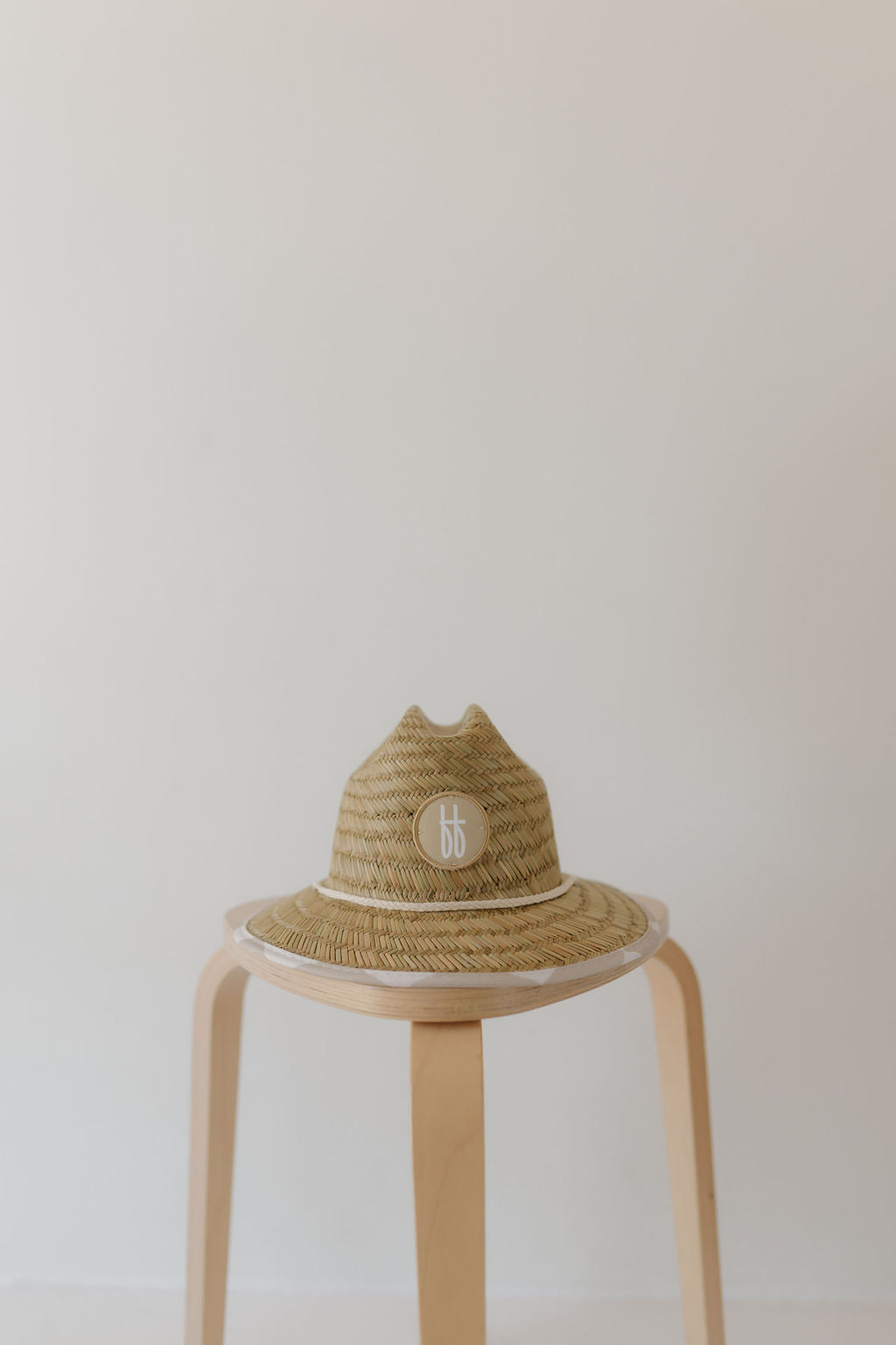 The Forever French Baby straw hat, ideal for beach days, displays a checkerboard pattern with a round logo on the front, elegantly set on a light wooden stool against a plain white background.