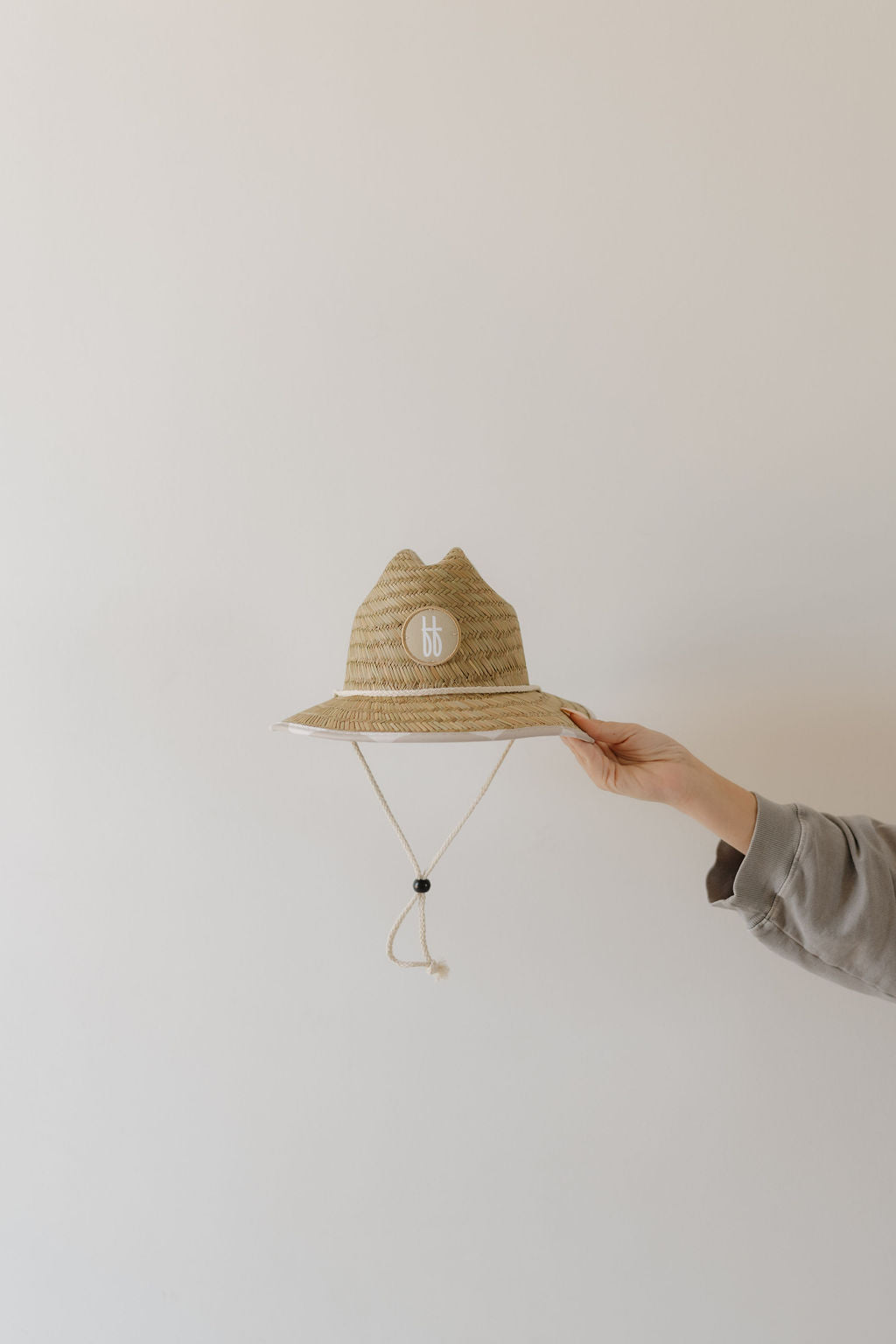 A hand holds the Straw Hat | Checkerboard by forever french baby against a plain background. The hat has a "bb" logo on the front and features a charming neutral checkerboard print with a chin strap.