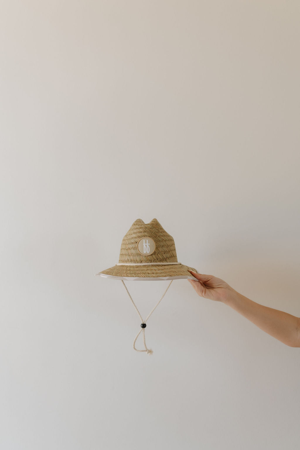 A hand holds the Straw Hat | Checkerboard, featuring a teardrop pinched crown and chin strap, against an off-white background. The hat has a small round "bb" logo on the front by forever french baby.