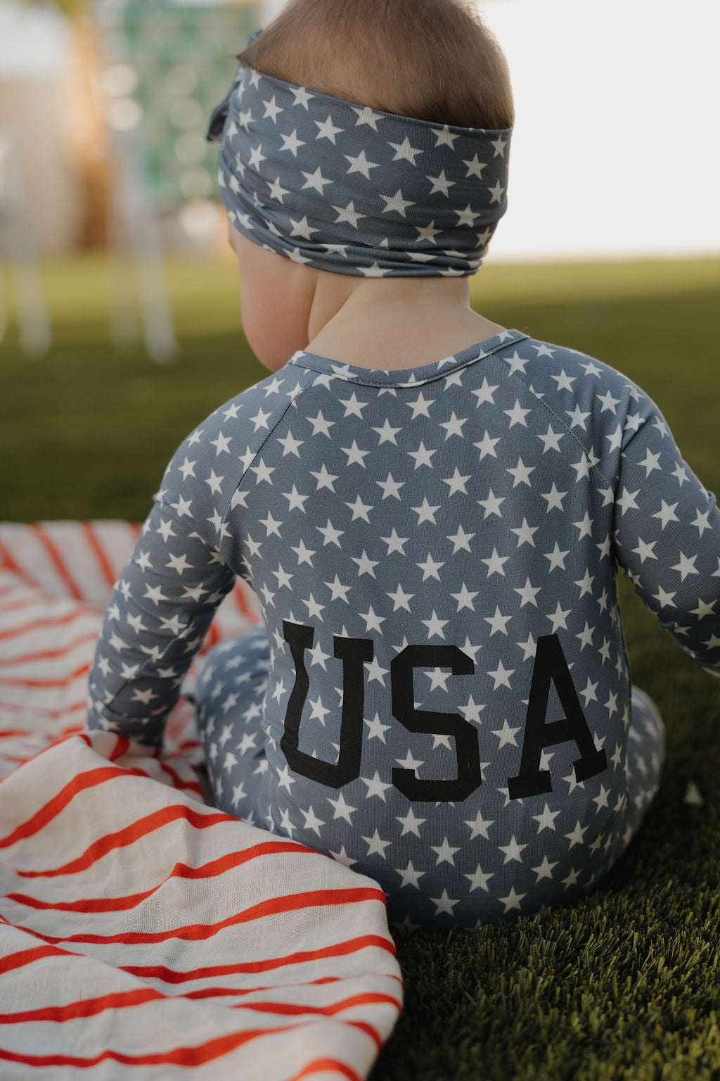 A young child sits on grass, facing away, in a blue outfit and the forever french baby Bamboo Headwrap | Star Spangled—an adjustable wrap with white stars and "USA" on the back. A red and white striped blanket lies nearby.