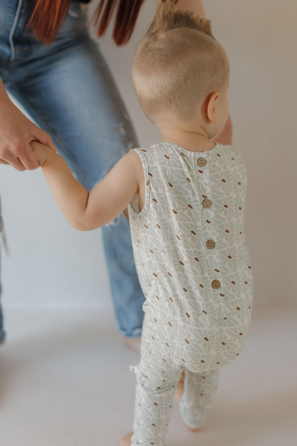 A toddler with a short mohawk wears the Harem Bamboo Romper by forever french baby, holding hands with an adult in blue jeans. Both stand indoors against a white background, enjoying hypoallergenic comfort.