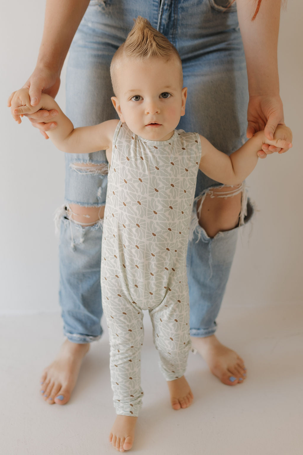 A toddler with a mohawk wears the fashionable Harem Bamboo Romper by forever french baby. They hold an adult's hands for support while the adult kneels in ripped jeans to assist. The indoor setting, with its light backdrop, beautifully complements the hypo-allergenic romper.