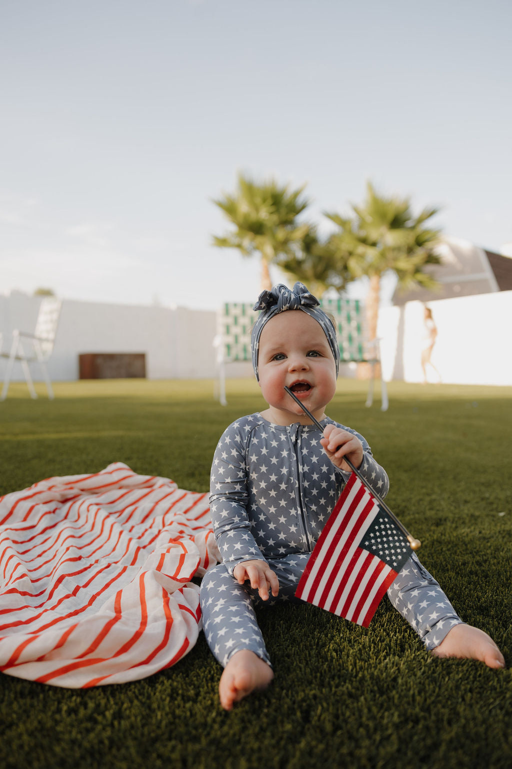 A baby wearing the forever french baby Bamboo Headwrap | Star Spangled sits on grass beside a striped blanket, holding an American flag, with palm trees and outdoor furniture in the background.