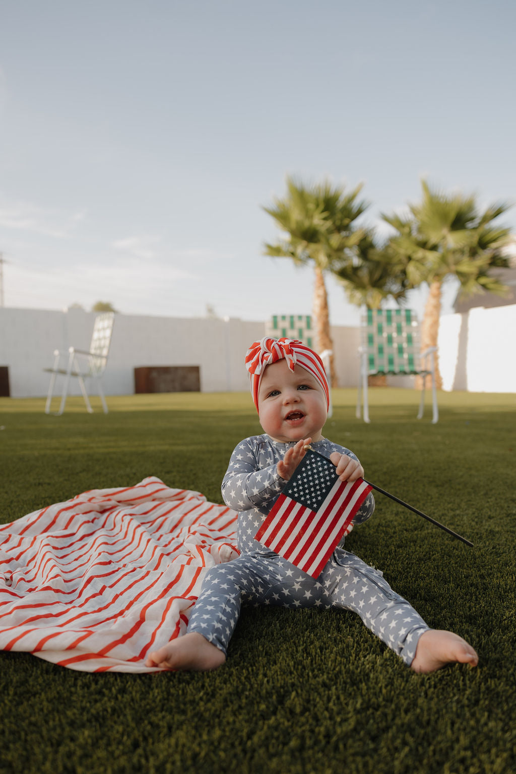 A baby in a star-patterned outfit and the forever french baby Bamboo Head Wrap | Stripes sits on grass, holding an American flag, with a striped blanket nearby and palm trees in the background.