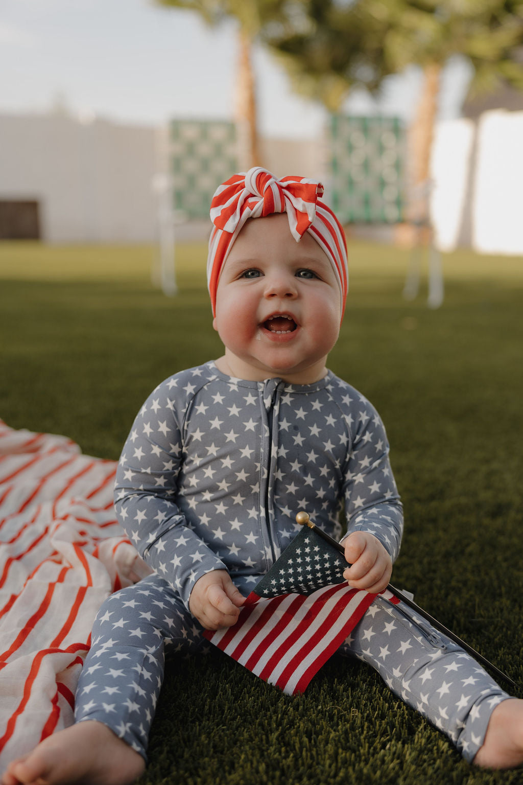 A smiling baby wearing a star-patterned outfit and a forever french baby Bamboo Head Wrap | Stripes sits on grass, holding a small American flag, with blurred trees and a white fence in the background.
