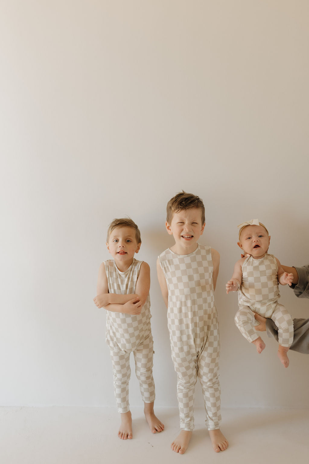 Three young children in matching Harem Bamboo Romper | Checkerboard by forever french baby stand against a beige background. Two older kids are barefoot side by side, while a baby is held by an adult, smiling and wearing a bow headband.