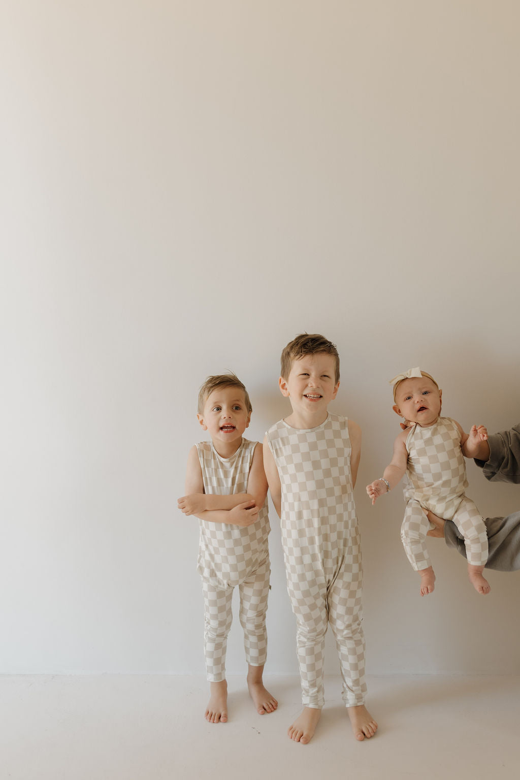 Three children pose against a plain background in matching forever french baby harem bamboo rompers with a light beige checkerboard pattern; the two boys have playful expressions, while the baby girl, wearing a bow headband, is held up by an unseen person.