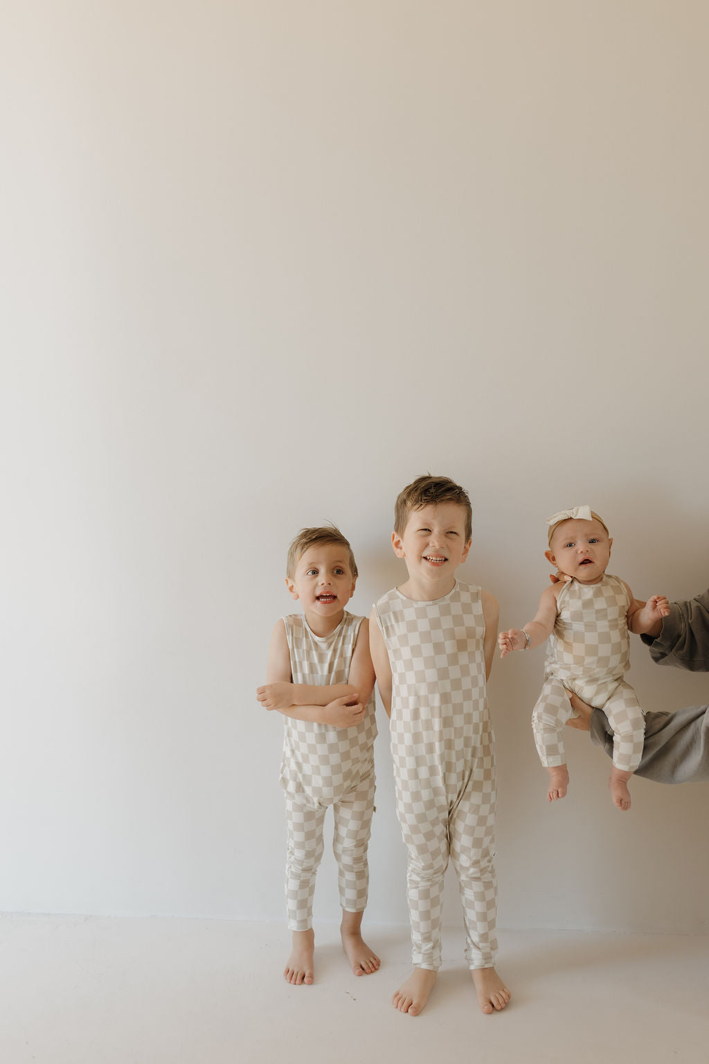 Three children in checkered outfits from forever french baby stand against a plain background. Two older kids are standing, and an adult holds a baby wearing the breathable Harem Bamboo Romper | Checkerboard. They are barefoot and smiling, creating a playful scene with their comfortable attire.