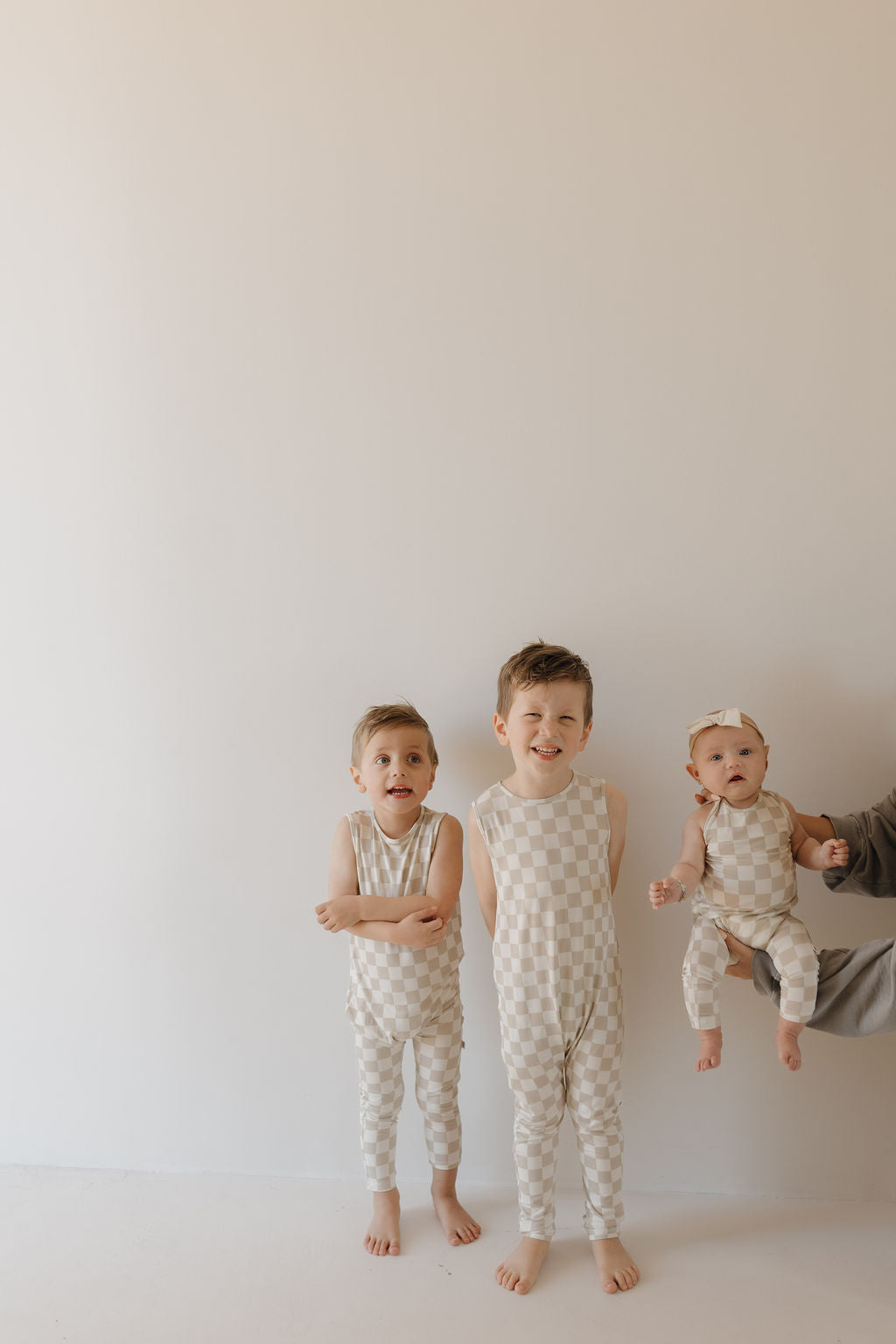 Three cheerful children stand indoors against a plain background. Two older kids wear matching forever french baby Harem Bamboo Rompers in a checkered design, with one making a funny face. A baby in similar breathable attire is held by an unseen person on the right.