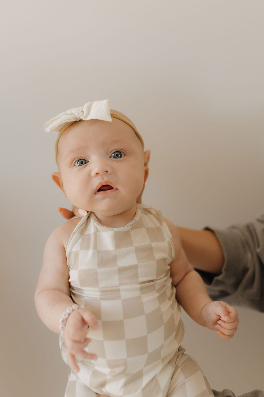 A baby in a breathable, light-colored "Harem Bamboo Romper | Checkerboard" by forever french baby and a headband with a bow is held by an adult's hand. Adorned with a bracelet, the little one gazes curiously at the camera against a plain background.