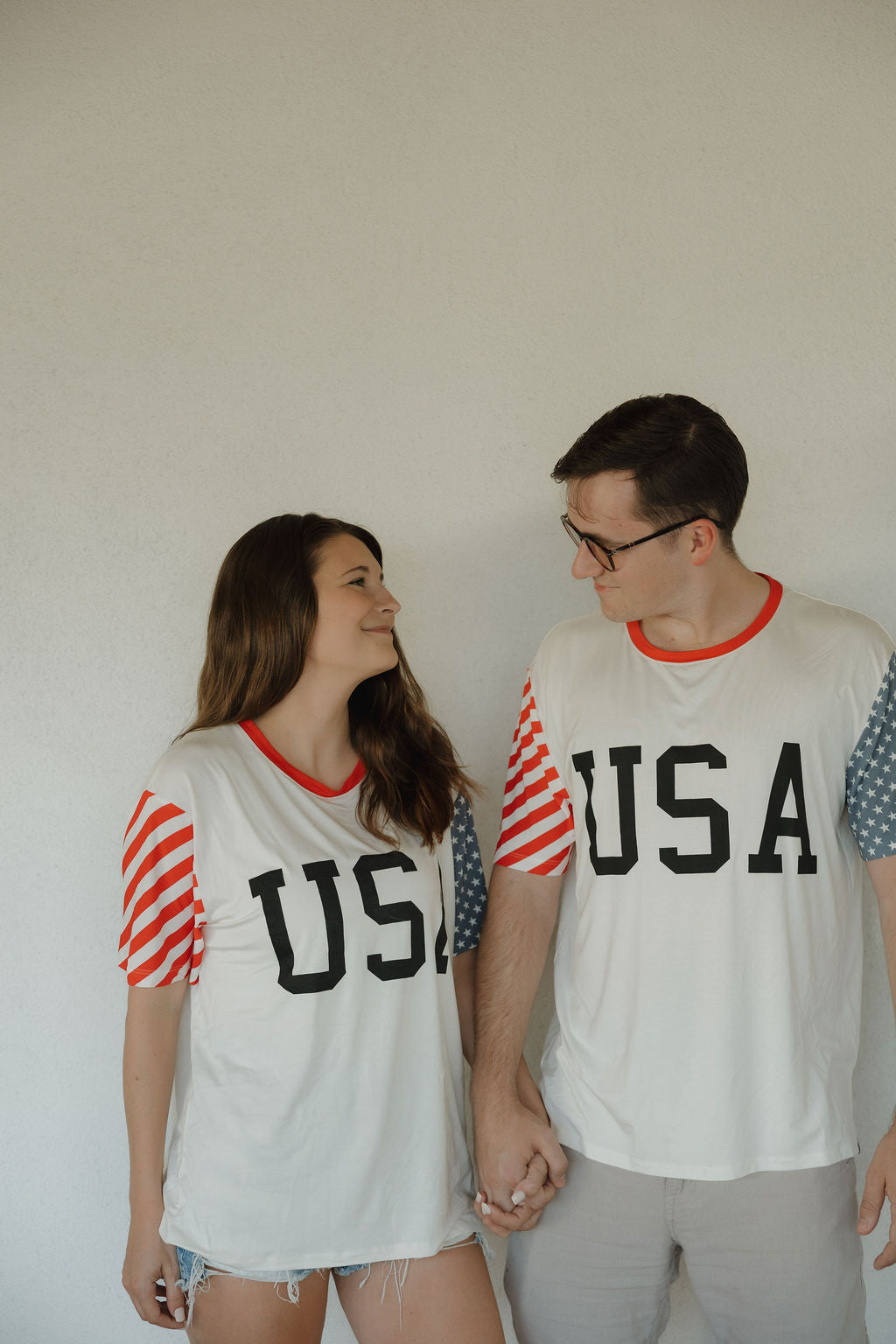 A man and woman hold hands, smiling at each other in matching forever french baby Adult Bamboo T-Shirts | Stars & Stripes, featuring "USA" on the front and red, white, and blue patterned sleeves against a plain light background.