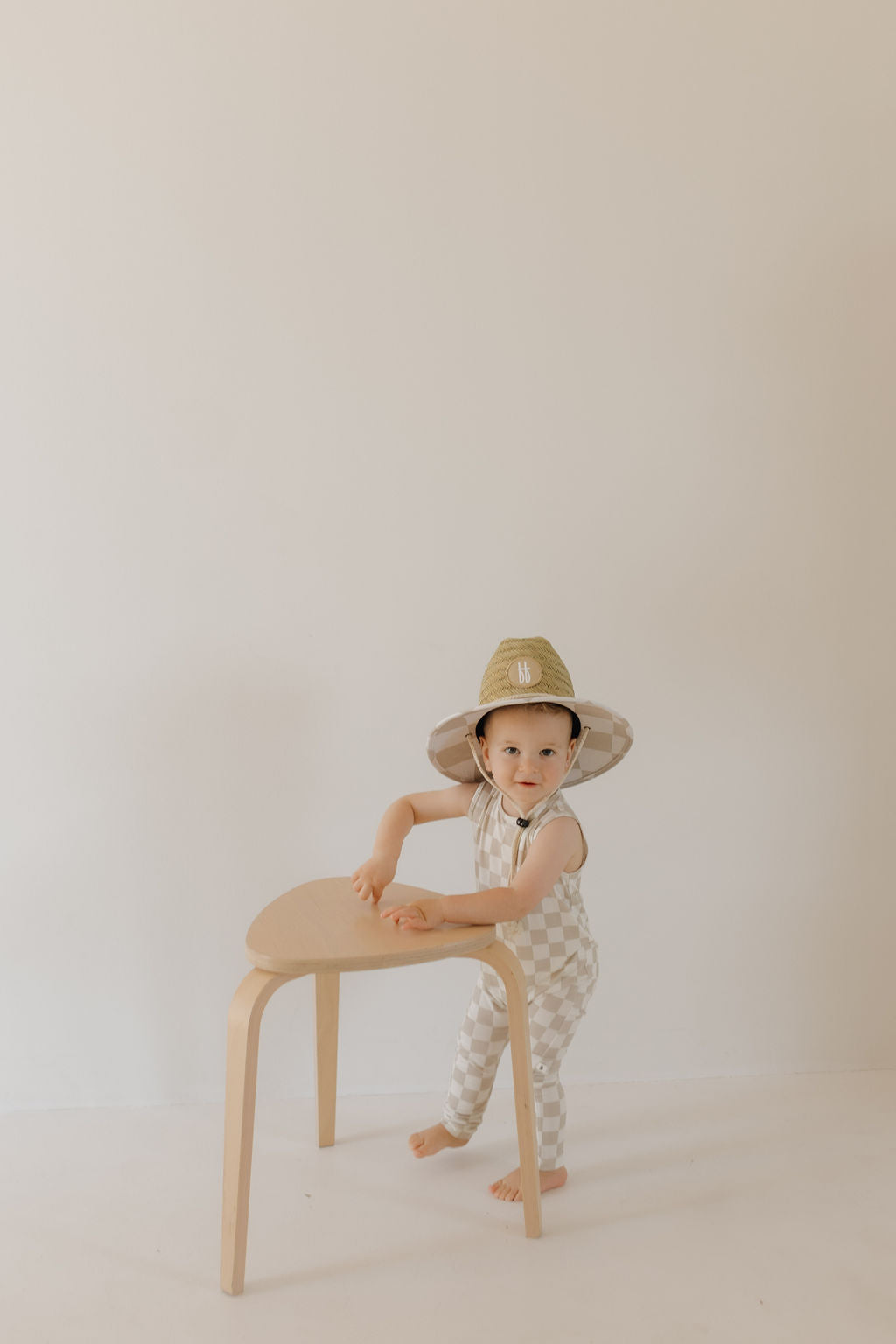 A toddler in a forever french baby Straw Hat | Checkerboard leans against a small wooden stool, with a minimal background emphasizing their gaze towards the camera against a neutral wall and floor.