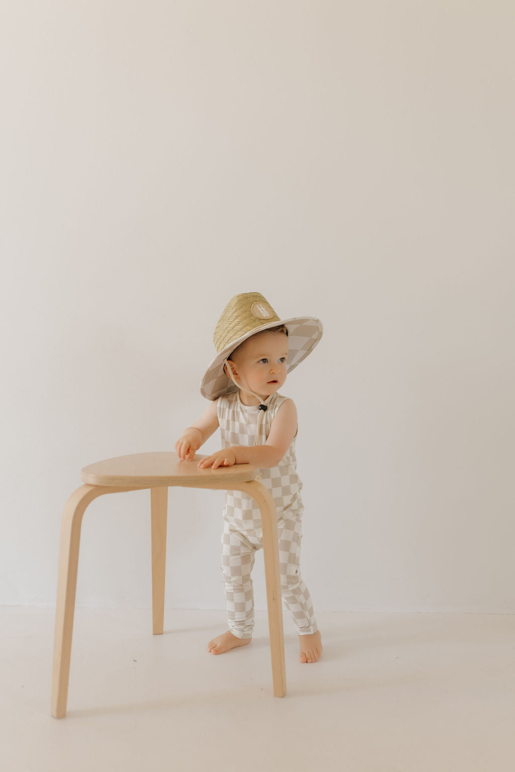 A toddler in a "Harem Bamboo Romper | Checkerboard" by forever french baby and a straw hat leans on a small wooden stool against a plain white backdrop.