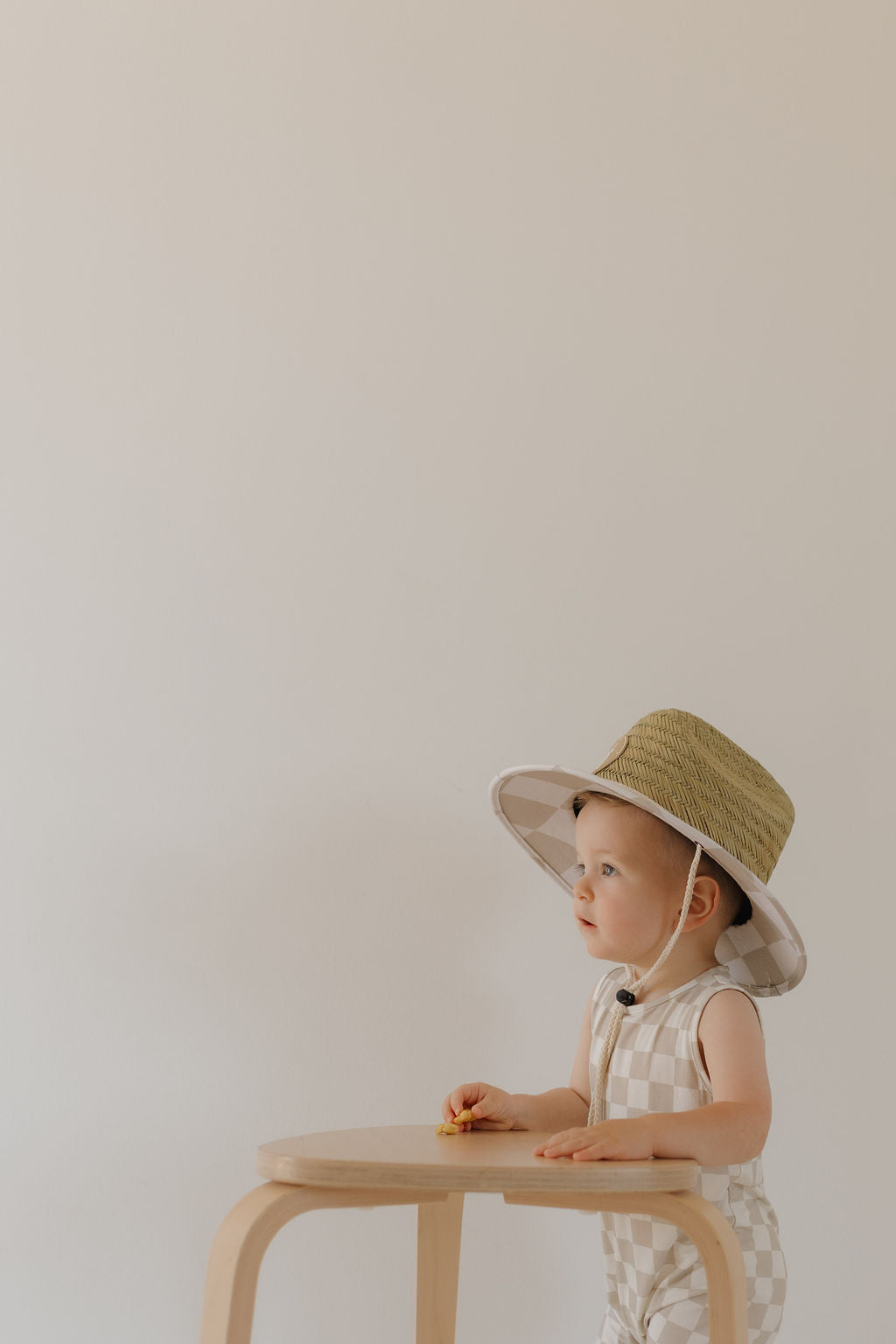 A toddler wearing forever french baby's Harem Bamboo Romper | Checkerboard and a straw hat sits at a light wooden table with a plain beige background.