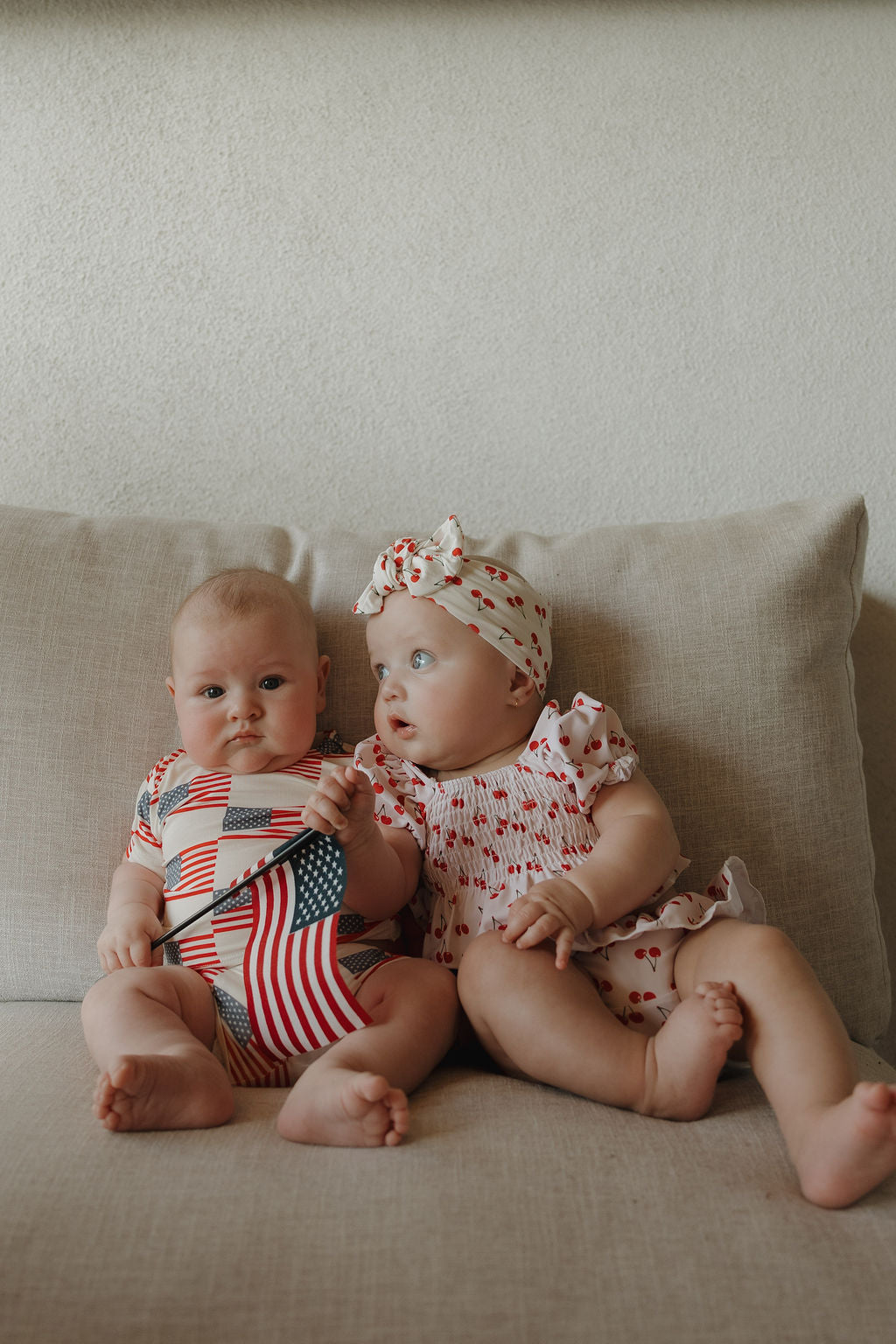 Two babies sit on a beige couch. The baby on the right wears the forever french baby Toddler Two Piece Swimsuit | Sweetheart Cherries, complete with matching headband, and looks at her friend in an American flag onesie.