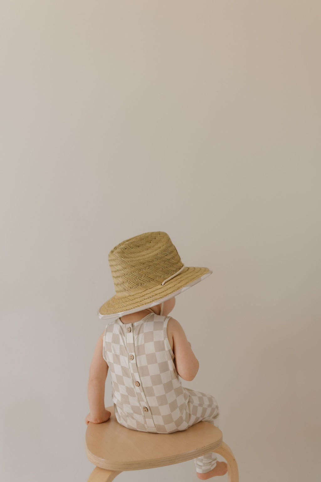 A child sits on a wooden stool facing away, dressed in Forever French Baby's checkerboard outfit and a wide-brimmed straw hat. The soft cream background evokes serene beach days, creating a minimalist and peaceful atmosphere.
