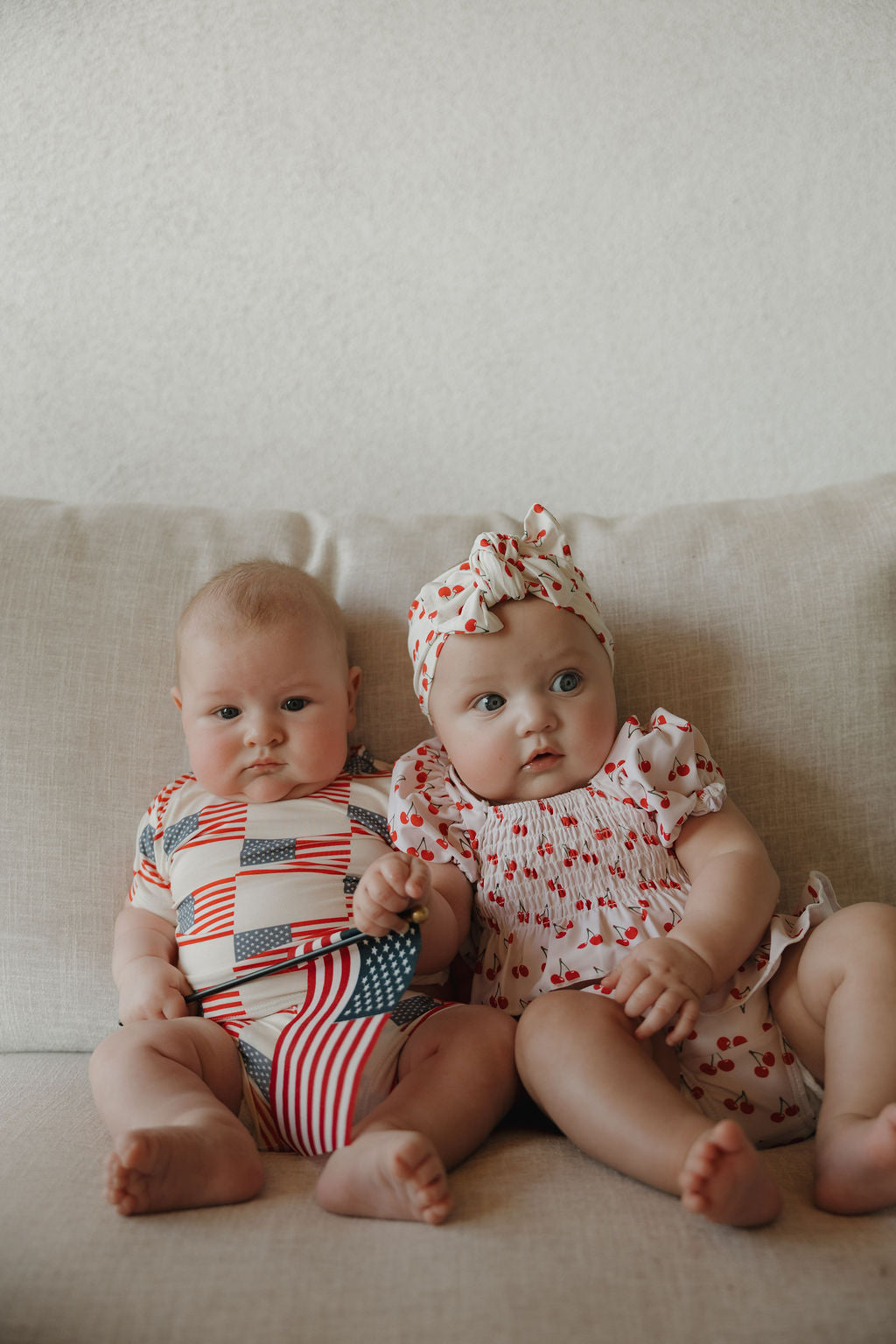 Two babies sit on a pale couch. The baby on the right wears the forever french baby Toddler Two Piece Swimsuit | Sweetheart Cherries, including a cherry print set with matching headband; the other wears a patriotic outfit.