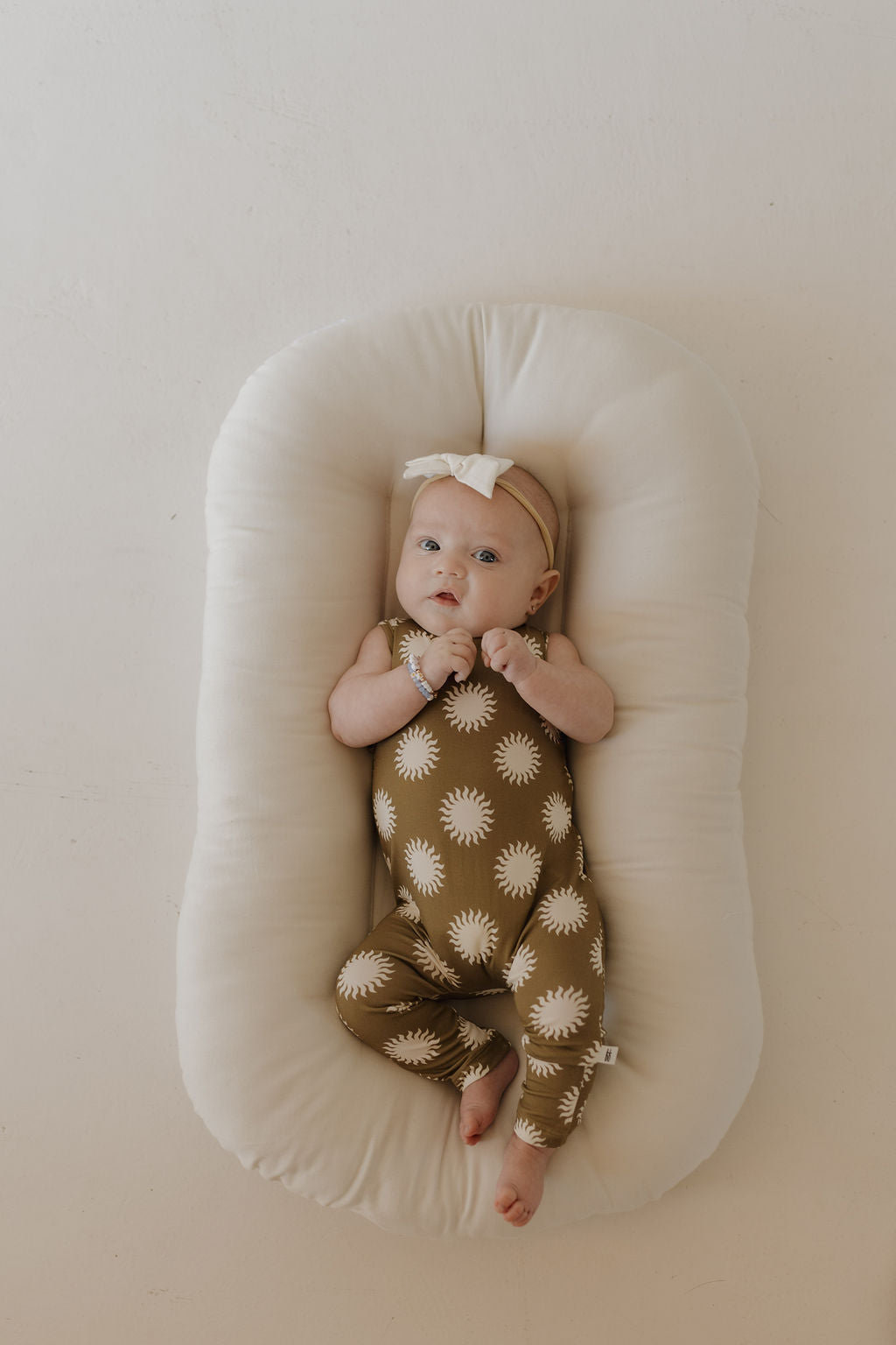 A baby wearing the "Harem Bamboo Romper | Sol" from forever french baby, featuring brown and white sun patterns in breathable bamboo, lies on a soft white pillow. The baby wears a beige headband with a white bow, gazing upward with hands near their mouth against a light neutral background.