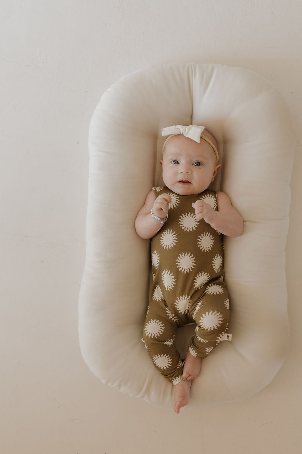 A baby rests on a soft, white cushion wearing the Harem Bamboo Romper | Sol by forever french baby, showcased with a matching bow headband. The breathable bamboo fabric offers comfort as the child gazes serenely against a neutral, light backdrop, adorned with white sun patterns.