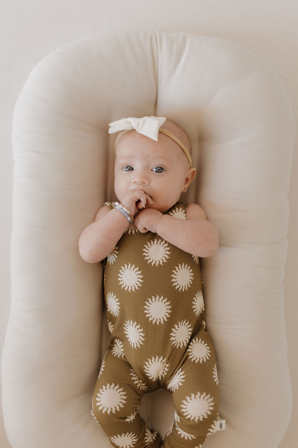 A baby in the forever french baby Harem Bamboo Romper | Sol lies on a soft, beige cushion. The brown onesie with white sun patterns, made from breathable bamboo fabric, ensures comfort and care. A bow-adorned headband completes the look against a minimal, soft-toned background.