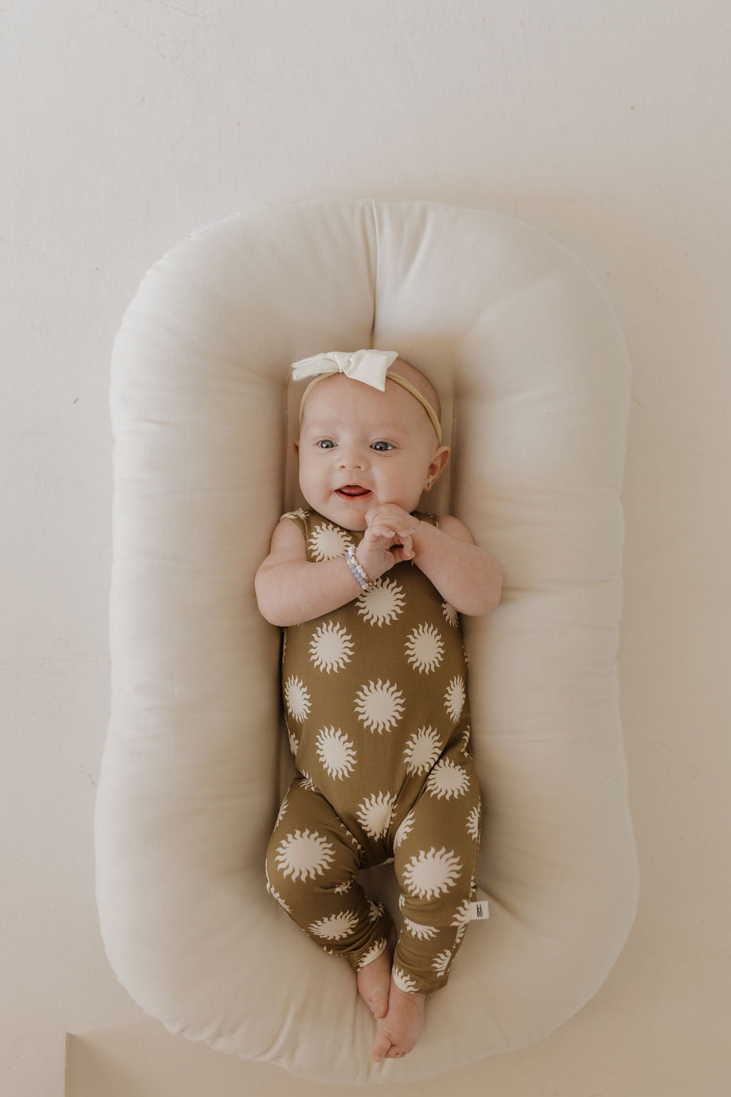 A baby in an olive-green Harem Bamboo Romper | Sol by forever french baby rests on a white cushion. Donning a light bow, the little one clasps their hands and gazes upward, perfectly comfortable in the breathable bamboo fabric.