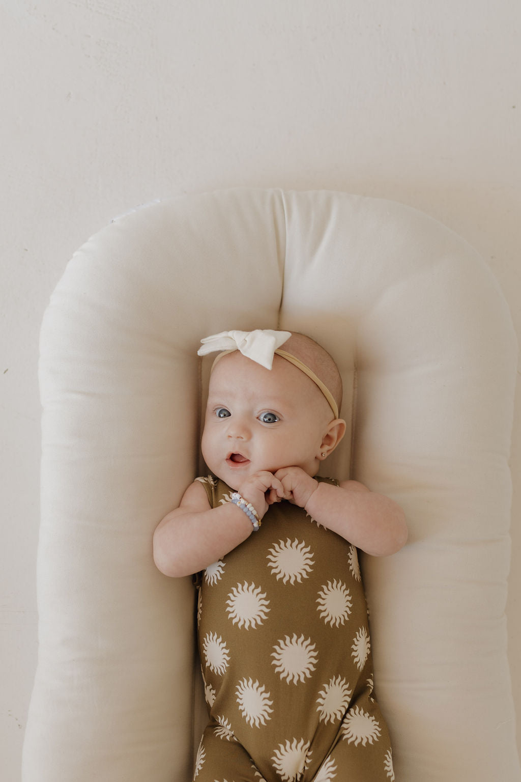 Wearing the "Harem Bamboo Romper | Sol" by forever french baby, a baby reclines on a white cushion. The green romper, adorned with white sun patterns and paired with a white bow headband, stands out against the neutral background as the baby curiously clasps their hands.