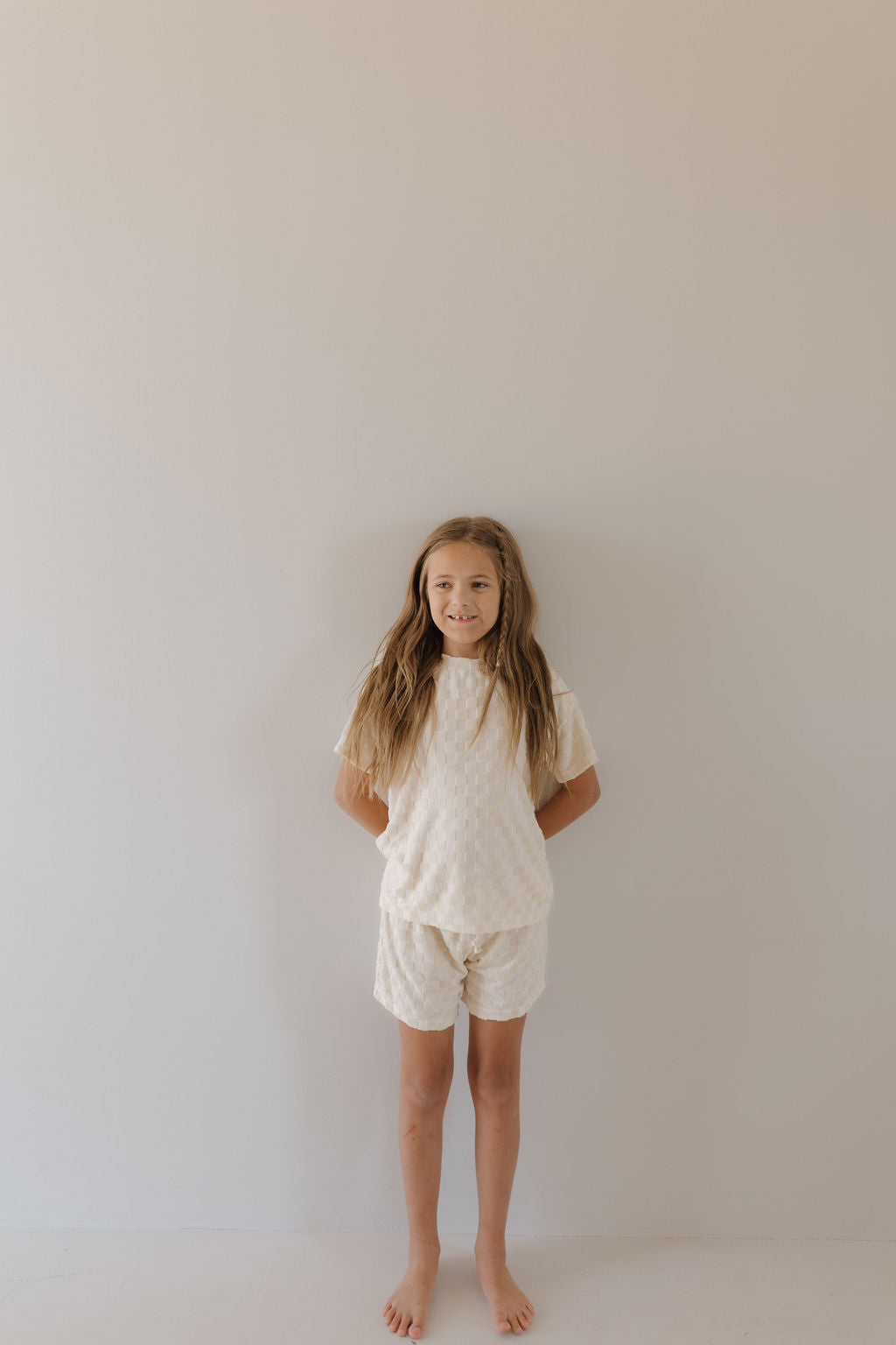 A young girl with long hair smiles against a plain light-colored wall, wearing the Terry Checkerboard Short Set in Cream by forever french baby. Her hands are behind her back and she is barefoot.