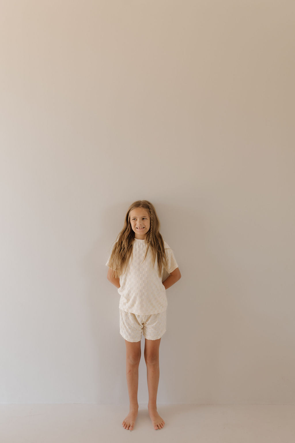 A girl with long hair stands barefoot against a plain beige background, wearing the Terry Checkerboard Short Set in Cream by forever french baby. She smiles coyly with her hands behind her back, perfectly embracing summer simplicity.