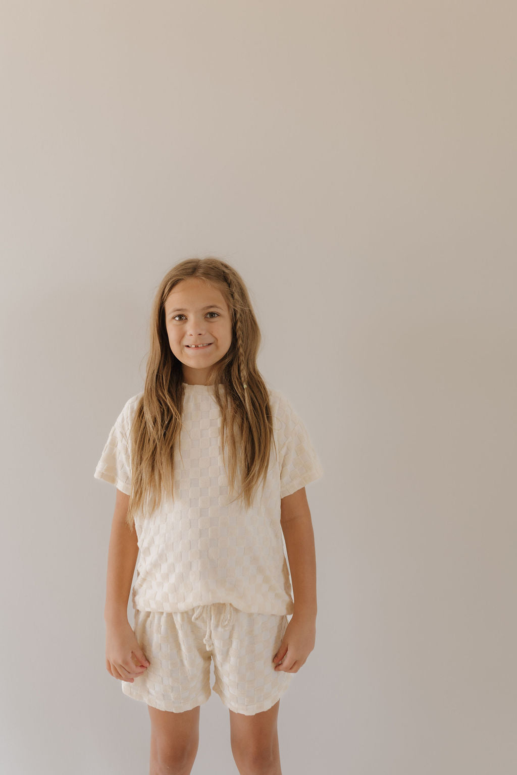 A young child with long hair stands against a light background, wearing a Terry Checkerboard Short Set in cream by forever french baby, smiling slightly.
