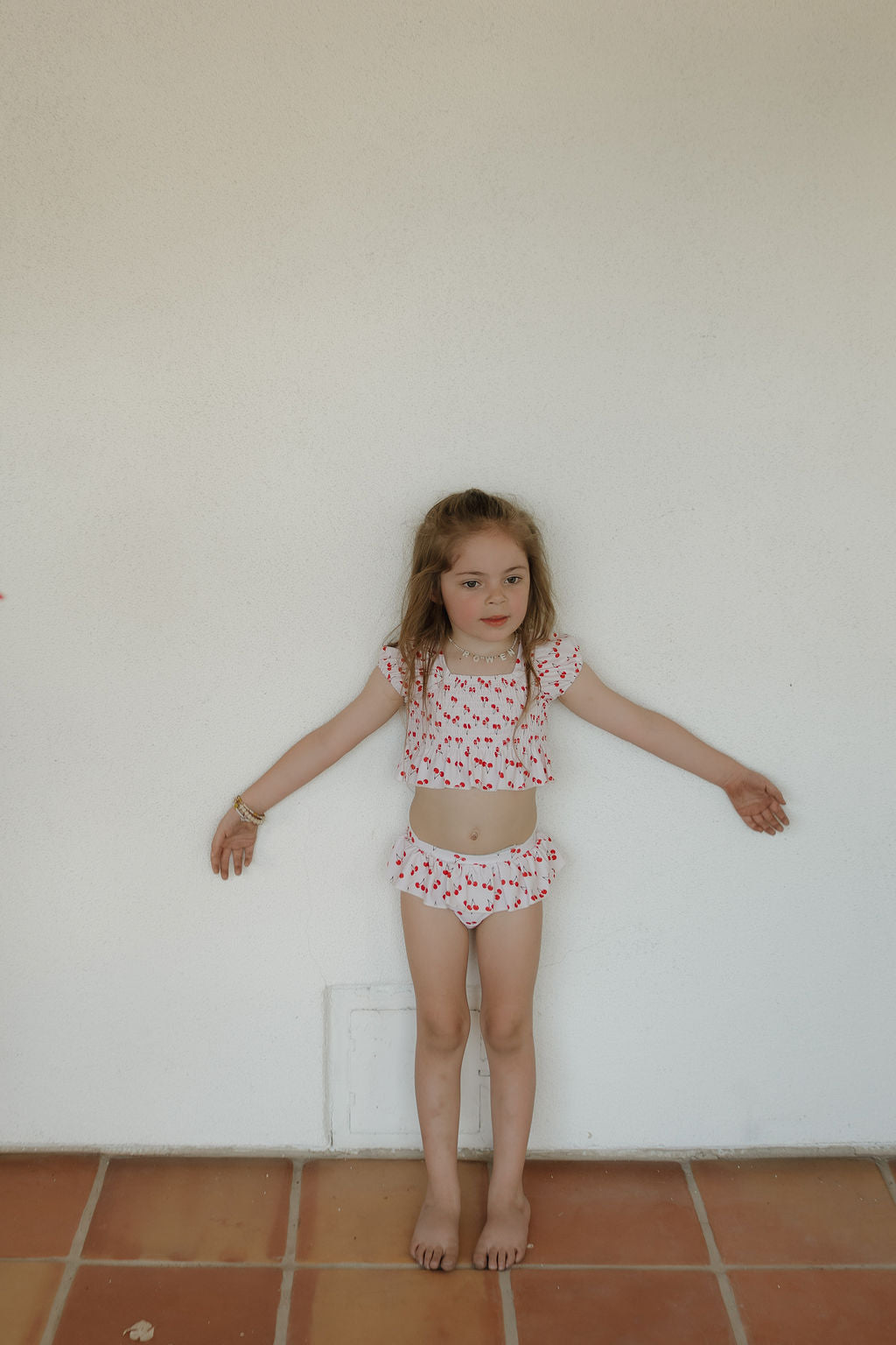 A young girl stands barefoot against a white wall, arms outstretched, wearing the forever french baby Toddler Two Piece Swimsuit | Sweetheart Cherries with ruffled sleeves and shorts.