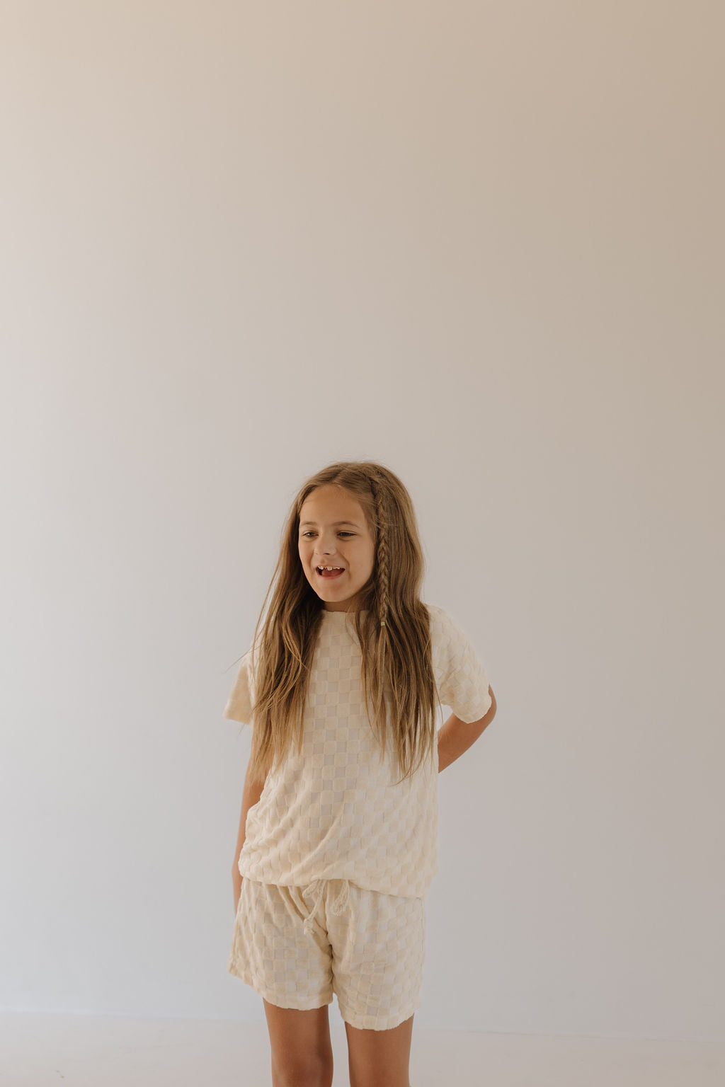 A young girl with long hair stands against a plain, light background wearing the Terry Checkerboard Short Set in Cream by forever french baby. Her hands are behind her back as she looks slightly to the side with a neutral expression, embodying chic summer style.