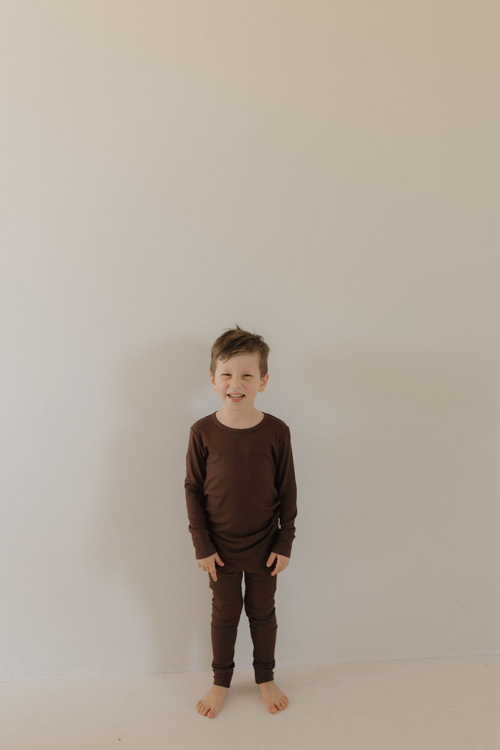 A young child with short hair smiles at the camera, barefoot against a plain wall, wearing the hypo-allergenic and gentle Bamboo Two Piece Lounge Set in Coffee Bean from forever french baby.