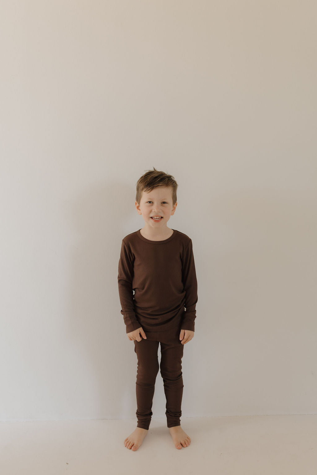 A young child with short hair stands barefoot against a plain light wall, smiling at the camera in the forever french baby Bamboo Two Piece Lounge Set | Coffee Bean—matching long-sleeve pajamas made from breathable bamboo fabric.
