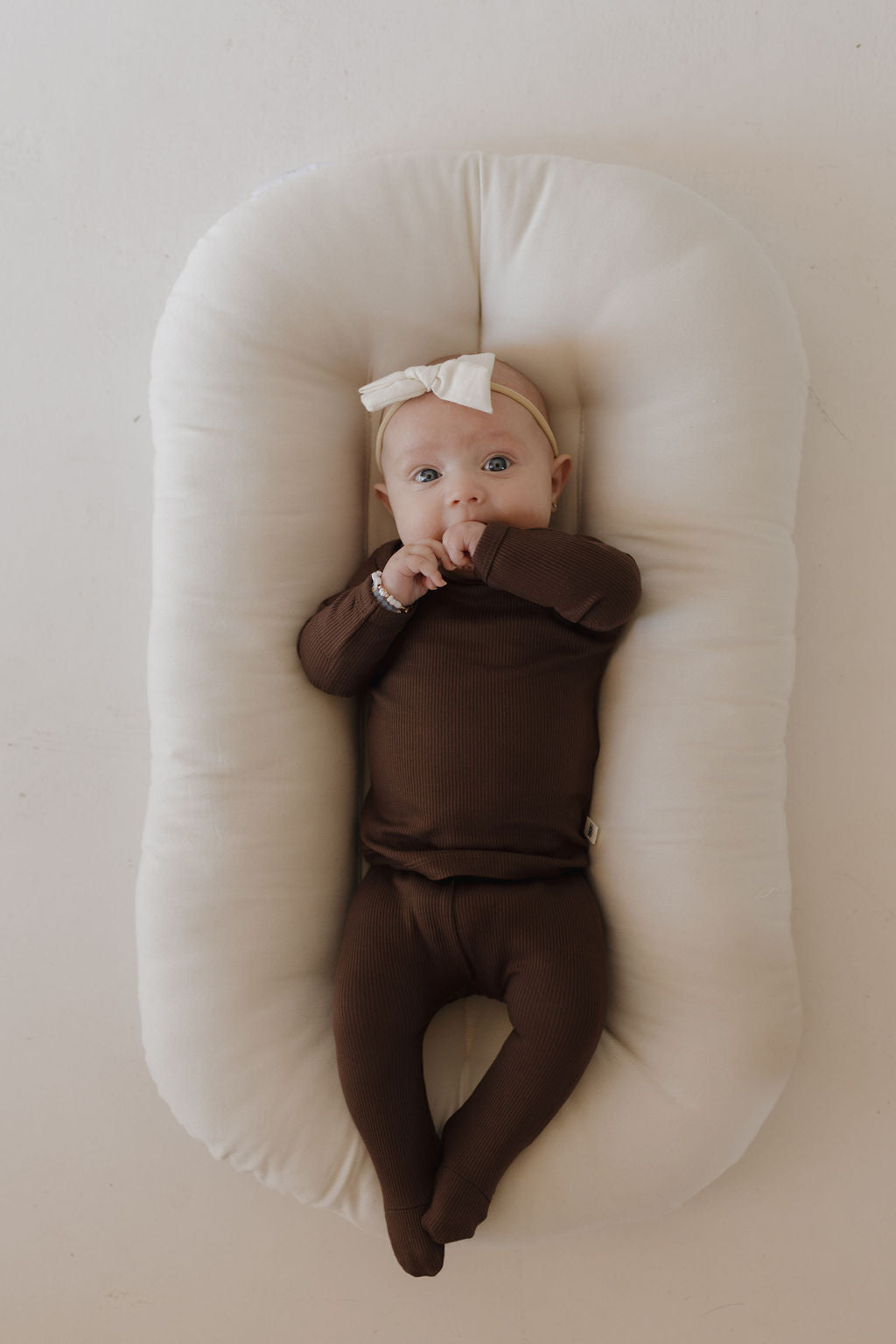 A baby dressed in the Sleepy Time Set | Coffee Bean by forever french baby and a cream bow headband lies on a cozy white cushioned pad. The hypoallergenic baby looks upward with a finger in their mouth, against a neutral, light background.
