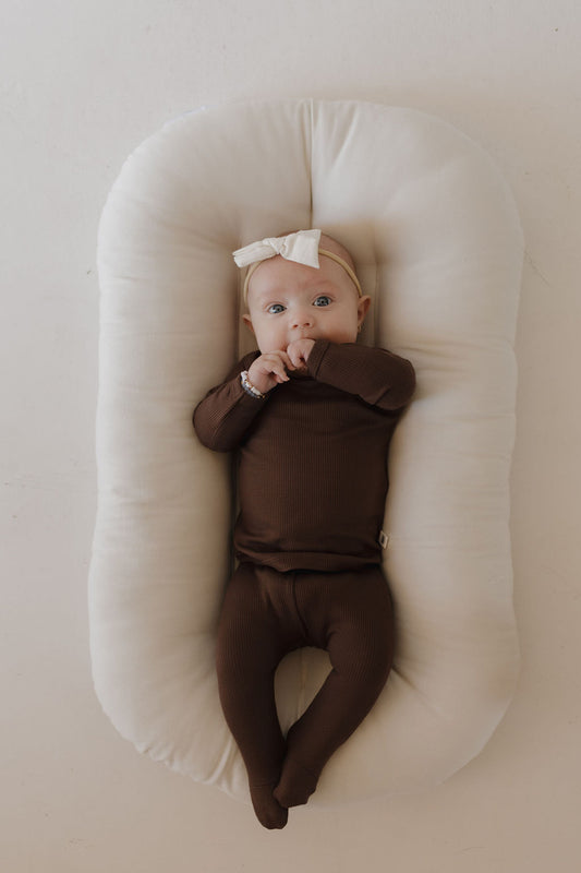 A baby dressed in the Sleepy Time Set | Coffee Bean by forever french baby and a cream bow headband lies on a cozy white cushioned pad. The hypoallergenic baby looks upward with a finger in their mouth, against a neutral, light background.
