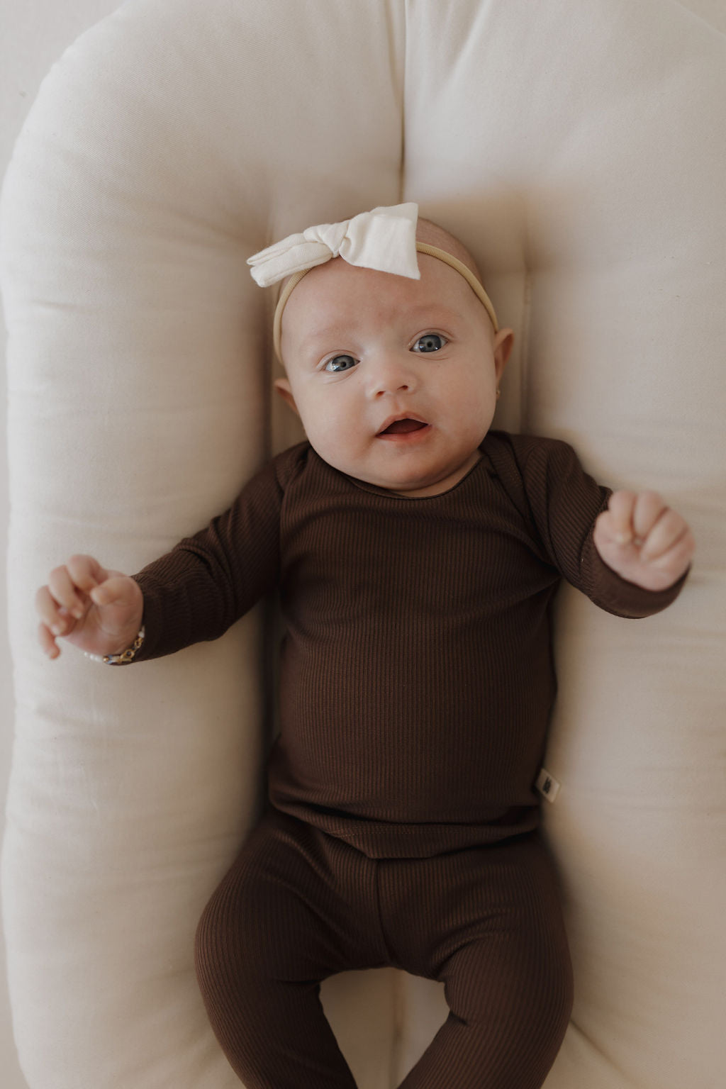 A baby in Forever French Baby's Sleepy Time Set | Coffee Bean lies on a cream cushion, wearing a breathable brown outfit and beige bow headband. The wide-eyed infant's raised arms enhance the charm, while the neutral background adds to the cozy, serene atmosphere.