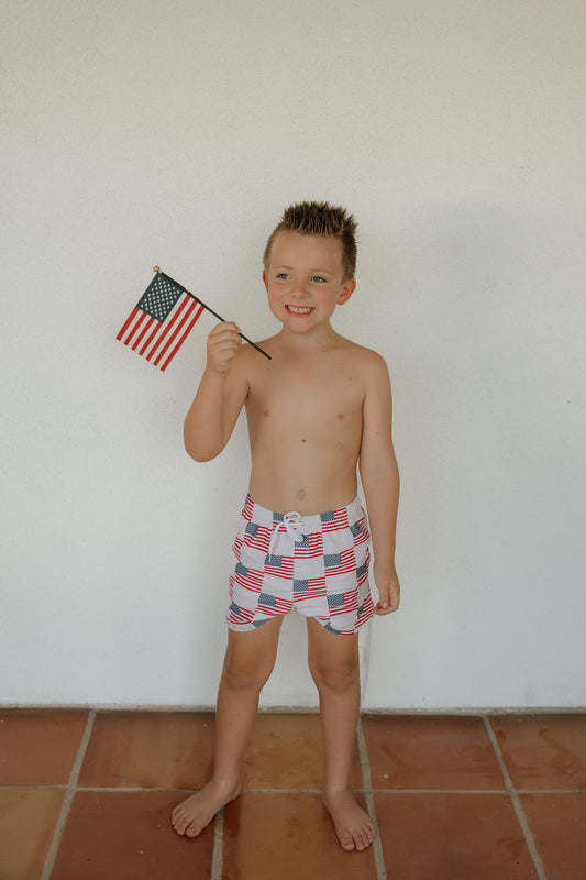 A smiling young boy wears the forever french baby Child Board Short | Americana with a functional drawstring, holding a small U.S. flag as he stands barefoot on a tiled floor against a plain white wall.