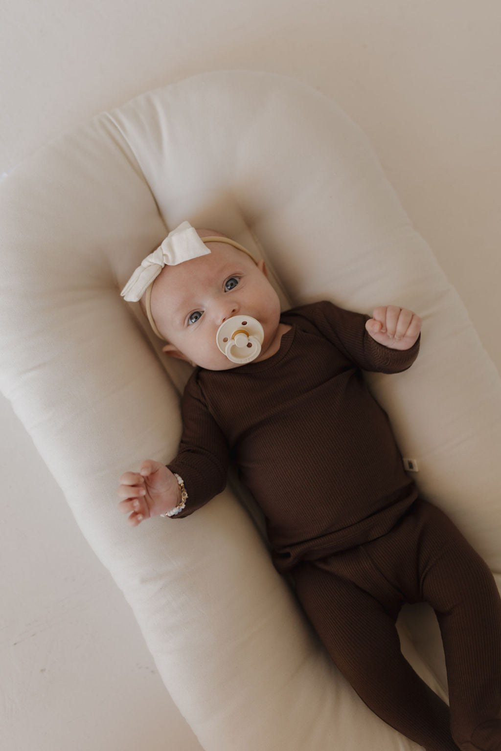 A baby cozily rests on a soft, white cushion wearing the Sleepy Time Set in Coffee Bean by Forever French Baby. The outfit is made of breathable bamboo and is paired with a pacifier and a white headband with a bow, all in softly lit surroundings.