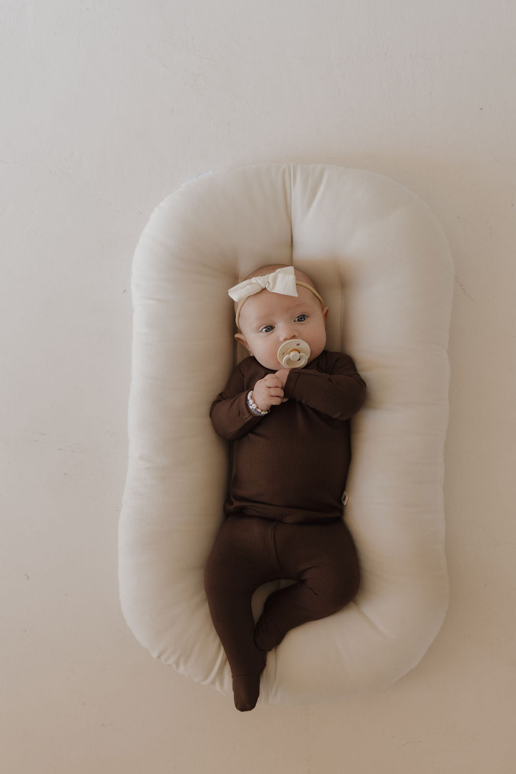 A baby in a breathable Sleepy Time Set | Coffee Bean from forever french baby lies on a soft white cushion holding a pacifier, with a small bow on their head. The background is neutral and light-colored.