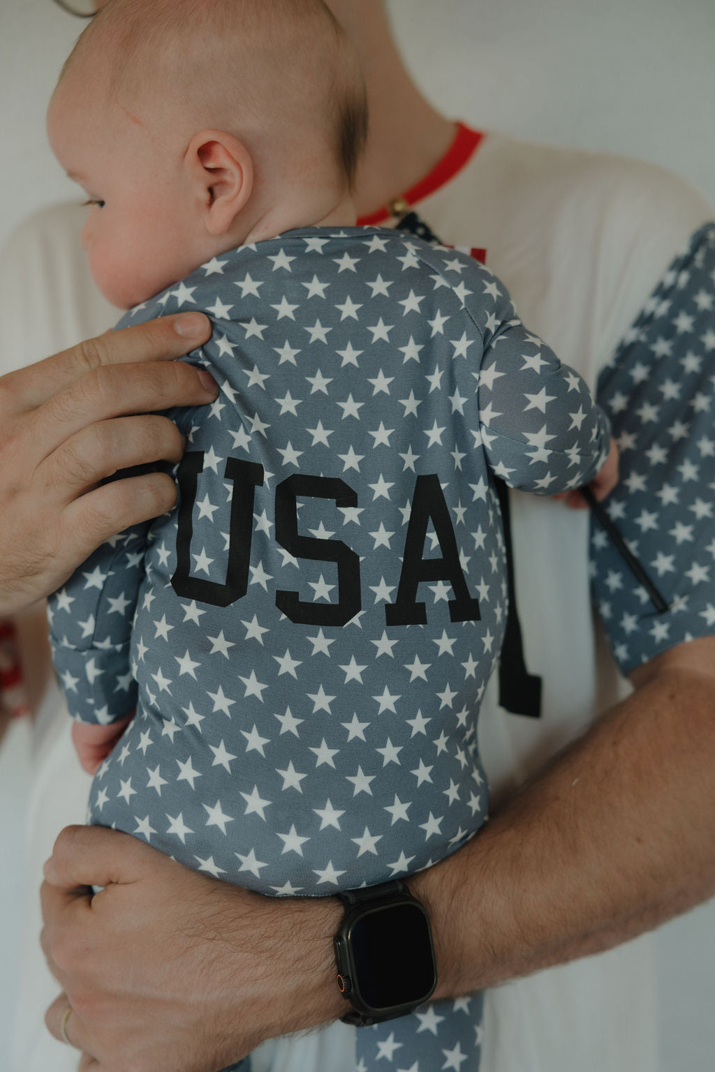 A person holds a baby dressed in forever french baby Bamboo Zip Pajamas | Star Spangled—a gray, breathable outfit with white stars and "USA" on the back. The person wears a smartwatch and a white shirt with red trim.