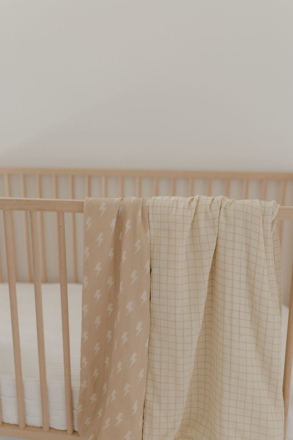 A wooden crib showcases two forever french baby muslin cotton blankets from the Muslin Quilt collection. One blanket features a tan and cream lightning bolt pattern, while the other has a checkered design, all set against a soft, neutral nursery backdrop.