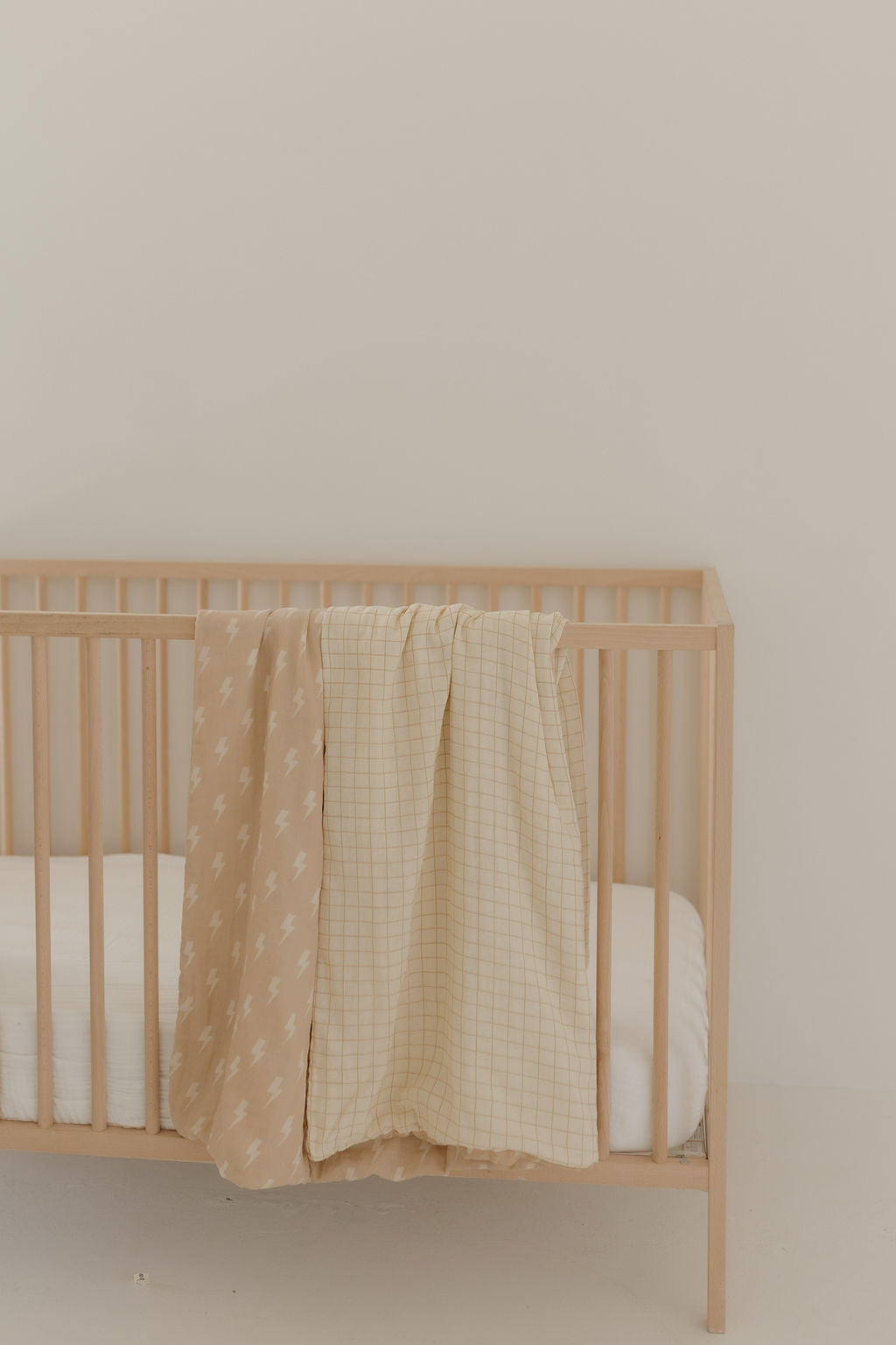 A wooden crib with a white mattress is shown, ideal for any baby nursery. Draped over the side are two soft muslin quilts—one featuring a beige pattern and the other a beige grid design, including the Muslin Quilt | MIS-PRINT Tan & Cream Lightning Bolt by forever french baby. A light-colored wall completes this serene setup.