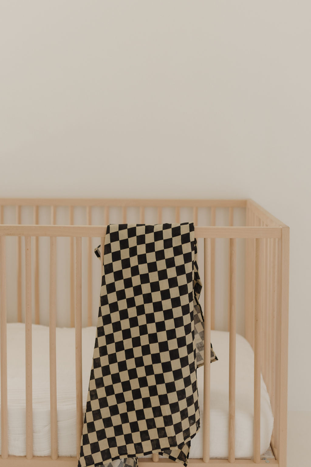 A light wooden crib with a white mattress features a forever french baby Muslin Swaddle | Black Checkerboard draped over the side, set against a plain off-white wall.