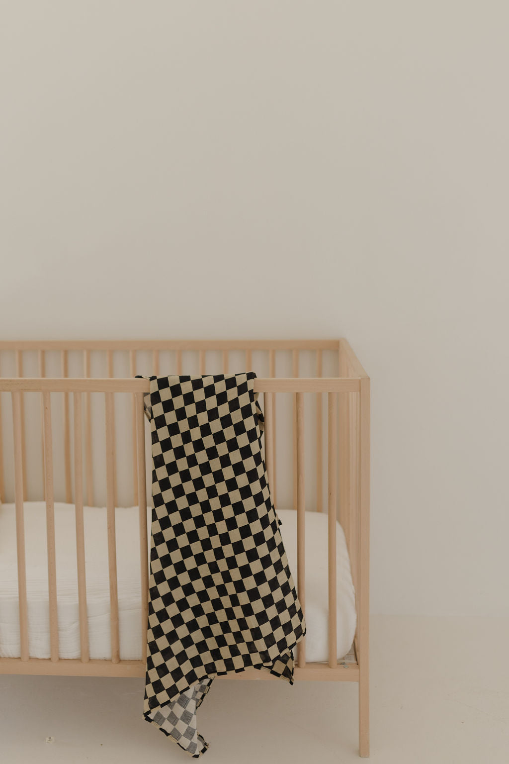 A light wooden crib with a white mattress stands against an off-white wall. Draped over the side is a forever french baby Muslin Swaddle | Black Checkerboard, adding a stylish touch with its black and beige checkerboard print.